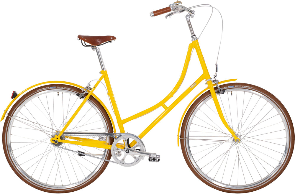 Cykel "Bike by Gubi Dame 7 Gear Gul" i gul farve, stående på asfalt med grønne træer i baggrunden.